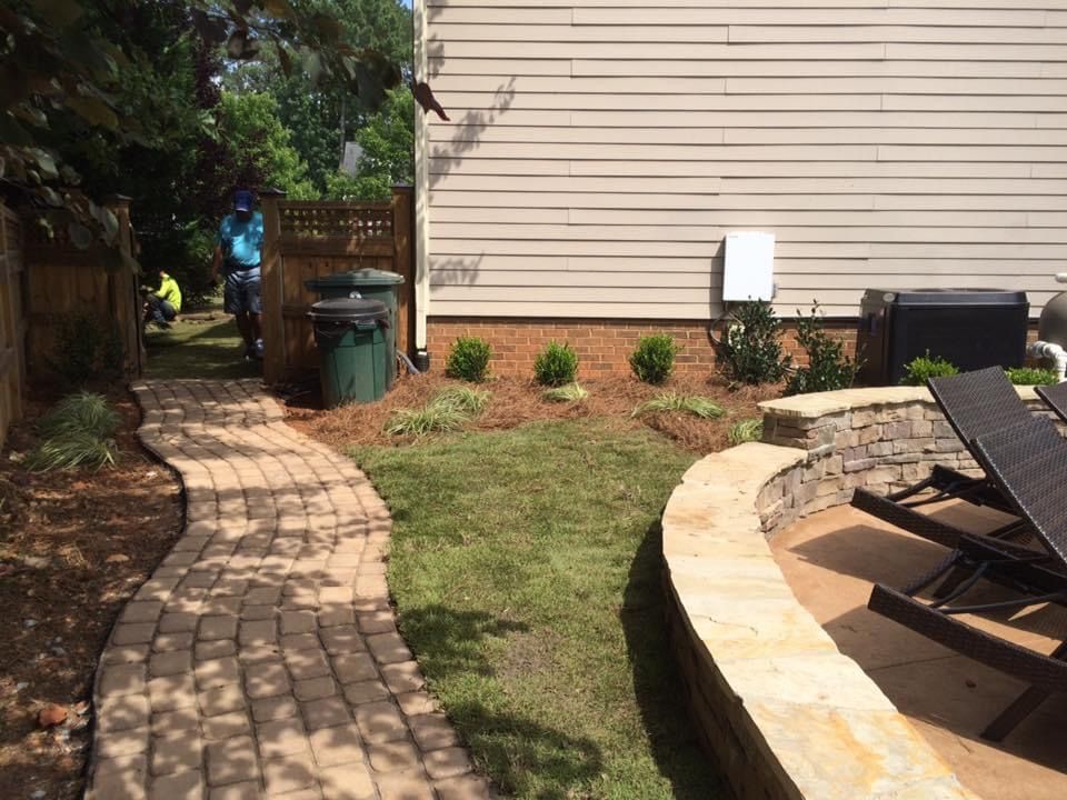 Brick garden path beside lawn and patio lounge chair, with trees, shrubs, and a trash bin near a house wall.