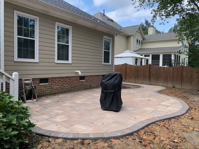 Backyard patio with stone pavers beside a house, curved border, fenced yard, and covered grill.