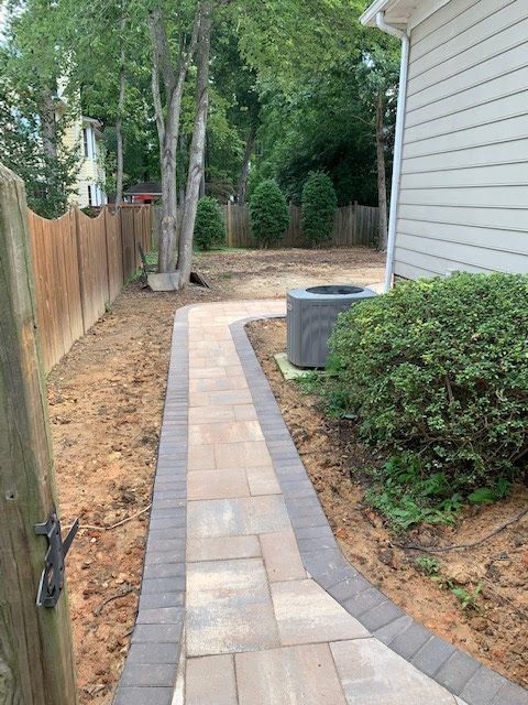 Narrow paved side yard path beside a house, with fence, trees, shrubs, and an AC unit.
