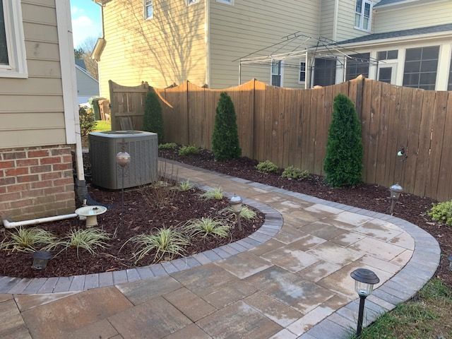 Curved stone patio path beside a house, wooden fence, shrubs, and an outdoor AC unit in a landscaped yard