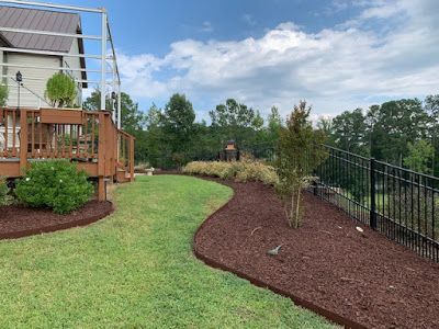 Curved backyard garden bed with mulch, shrubs, and a green lawn beside a wooden deck and black fence.