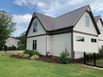 White house with a gray metal roof and black fence, set in a green yard under a partly cloudy sky