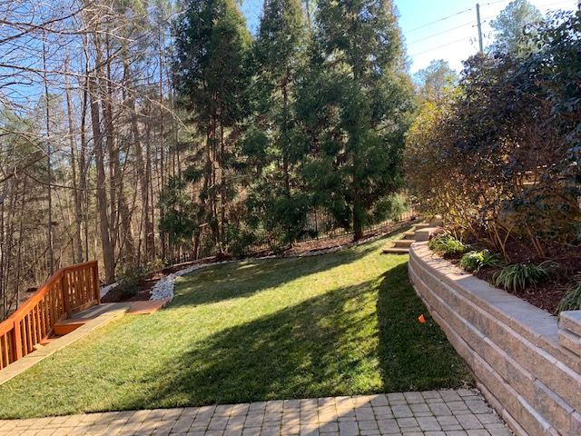 Backyard lawn with stone retaining wall, wooden deck, and tall trees in sunlight