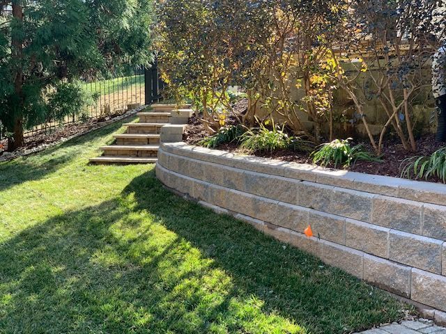 Backyard garden with stone retaining wall, steps, and sunlit grass under trees