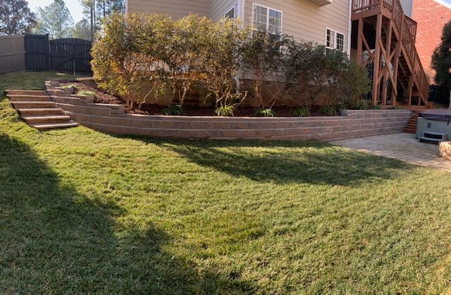 Grassy backyard with stone retaining wall, shrubs, stairs, and a house deck in the background