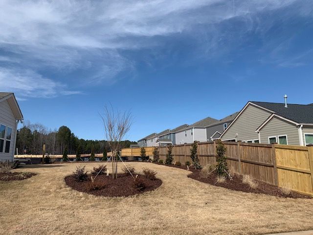 Backyard with a circular mulch bed, tan grass, wooden fence, and houses under a blue sky.