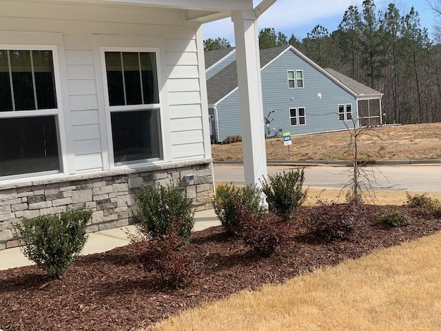 Front porch with white siding, small shrubs, and mulched beds, overlooking a suburban house and driveway.