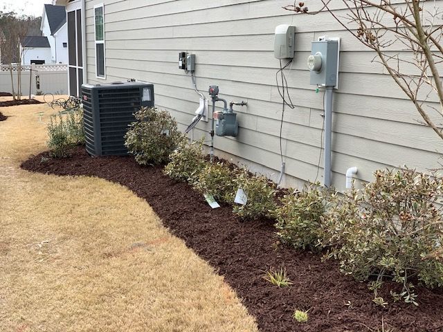 Side yard with shrubs and mulch along a house wall, beside utility meters and an outdoor HVAC unit