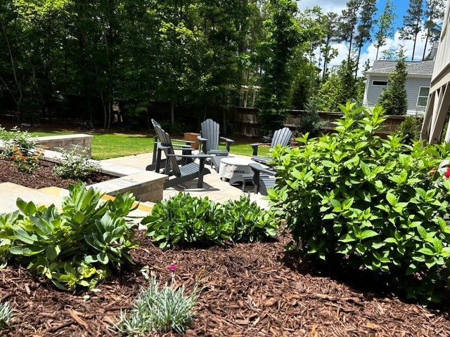 Backyard patio with black chairs and fire pit, surrounded by green shrubs and trees