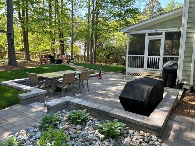 Backyard patio with outdoor dining table, grill, seating, and a screened porch amid trees and stone pavers