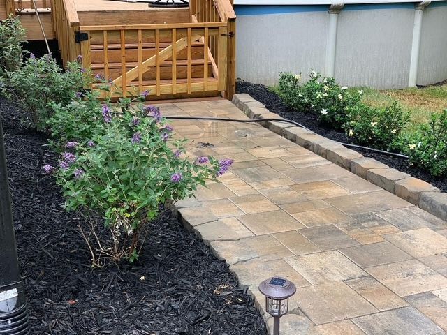 Garden walkway with tan pavers, black mulch beds, shrubs, and a wooden gate at the end
