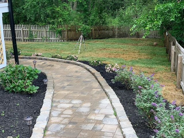 Curved stone garden path beside flower beds, with a grassy yard and wooden fence in the background