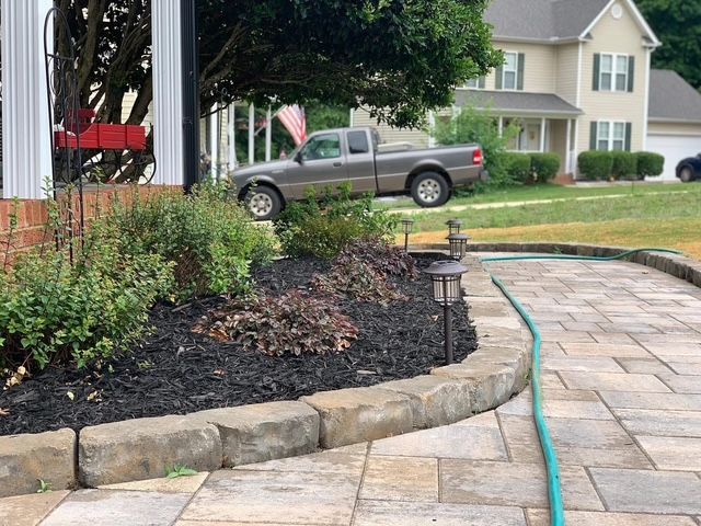 Curved stone garden bed beside a paver driveway with a green hose and a parked SUV in the background