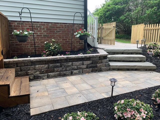 Backyard patio with stone steps, brick retaining wall, pavers, and flower beds leading to a fenced gate