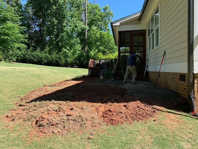 Workers spreading red dirt beside a house under construction, with a grassy yard and trees nearby