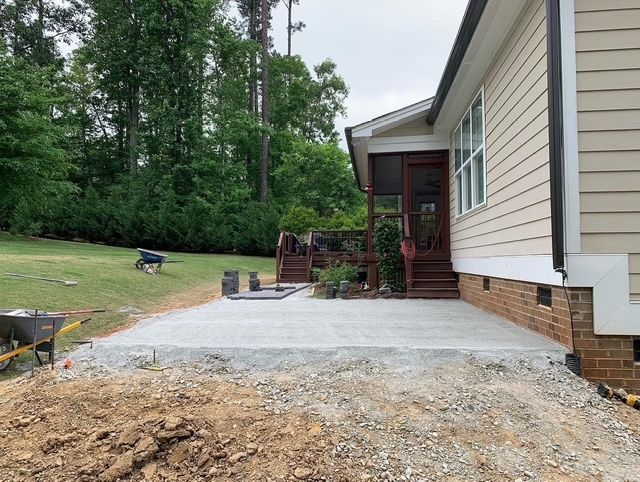 Concrete patio beside beige house with covered porch, surrounded by yard and trees