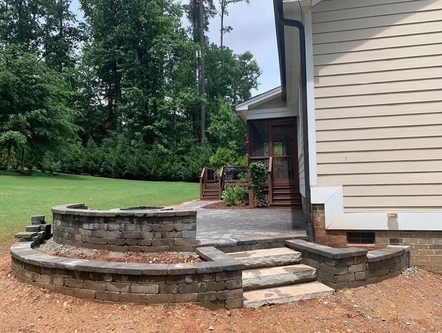 Backyard stone patio with curved retaining wall, steps, and beige house siding amid green lawn and trees