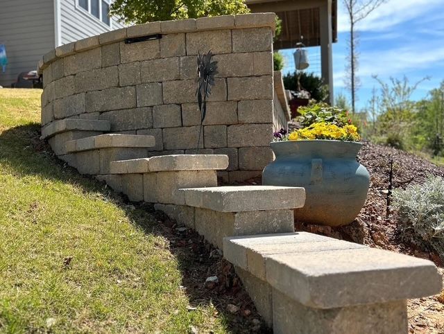 Curved stone retaining wall with steps beside a grassy slope and a blue planter with yellow flowers
