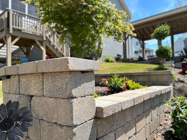 Stone retaining wall with flower bed, stairs, and a house deck in a sunny yard