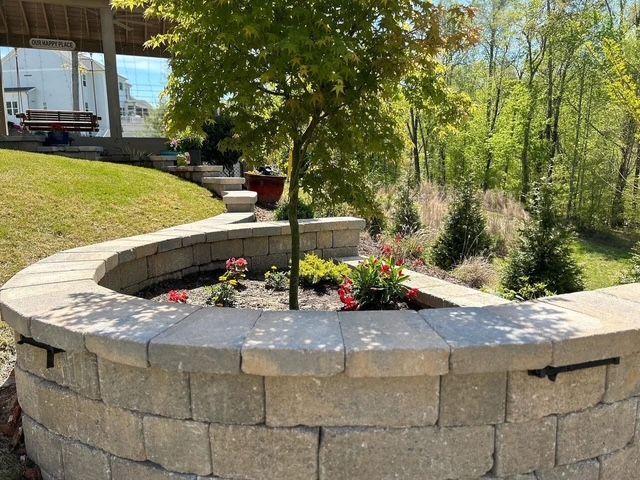Curved stone planter wall with flowers beside a grassy yard and wooded garden path