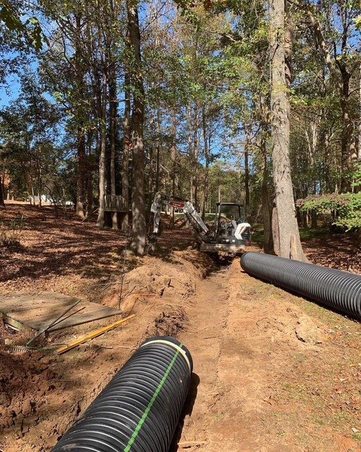Workers installing black drainage pipes on a wooded dirt slope.