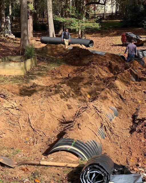 Workers install a large black pipe in a dirt trench at a wooded construction site.