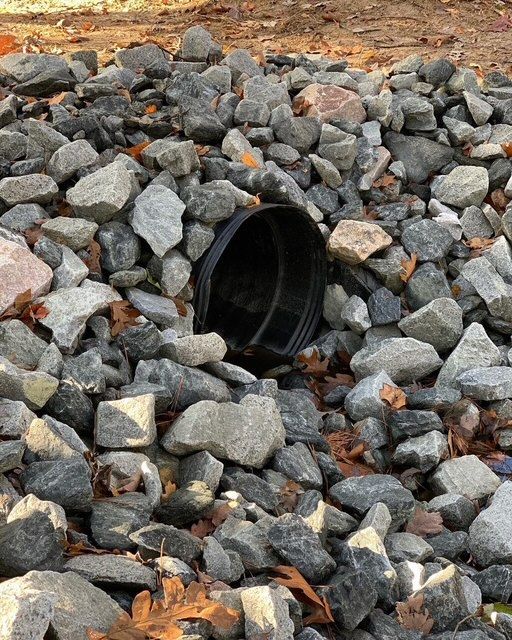 Black drainage pipe opening surrounded by gray rocks and orange leaves outdoors