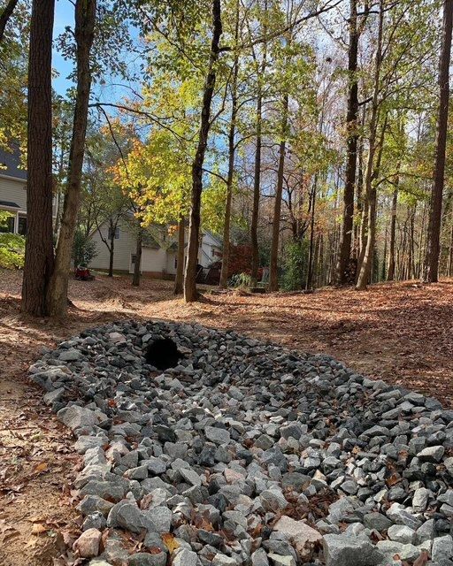 Rock-lined drainage ditch beside a wooded yard and house in autumn.