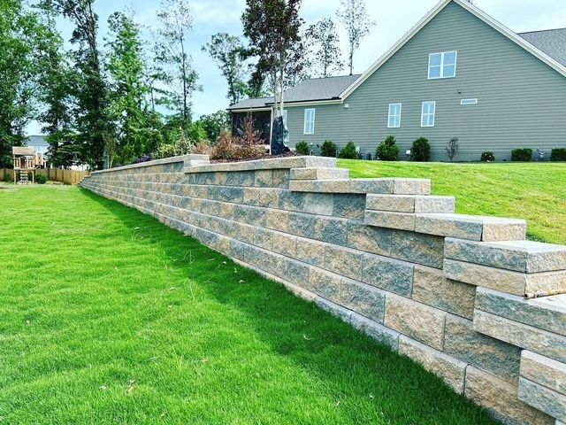 Stone retaining wall beside a green lawn, with a house and trees in the background.