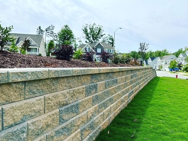 Stone retaining wall along a grassy slope in a suburban neighborhood with houses and trees behind it