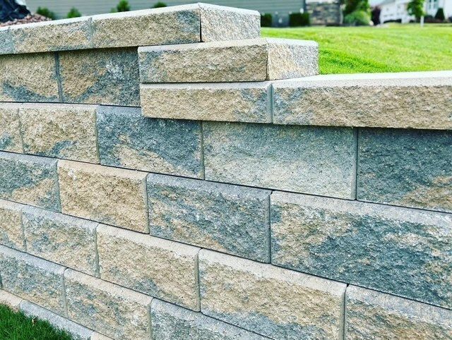 Staggered gray stone retaining wall with tan capstones beside a grassy lawn.