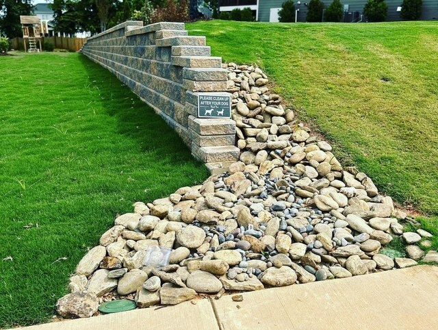 Stone drainage wall with a rock spillway on a grassy slope beside a sidewalk