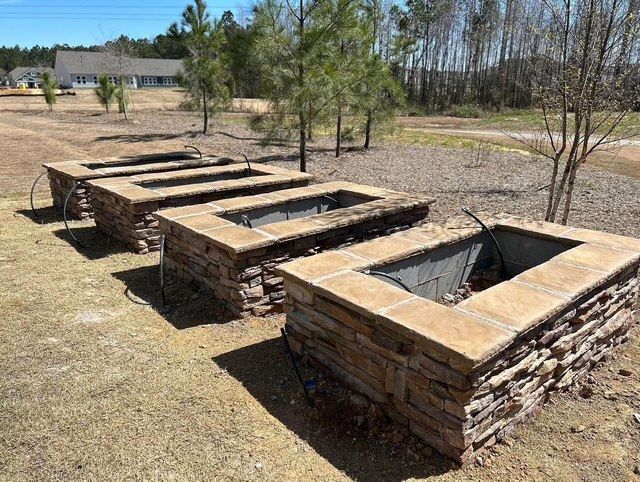 Three stone fire pits in a sunny outdoor park area with sparse trees and dry grass