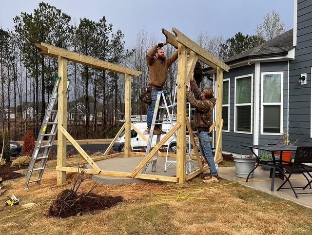 Two people assembling a wooden frame beside a house, with ladders and tools in a yard.