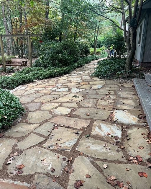 Stone garden path curving through lush greenery beside a building, with a few people in the distance.