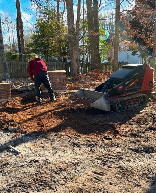 Person using a compact loader to move dirt near stacked bricks in a wooded yard