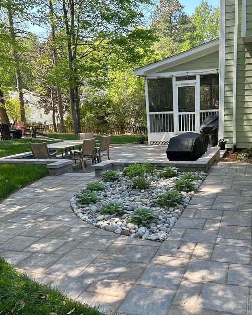 Backyard patio with stone pavers, a curved garden bed, outdoor seating, and a grill beside the house.