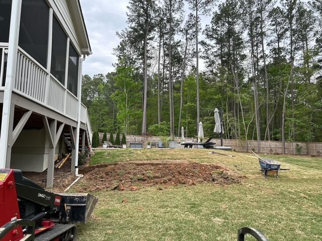 Backyard with raised deck, bare soil mound, wheelbarrow, patio chairs, and tall trees beyond a wooden fence