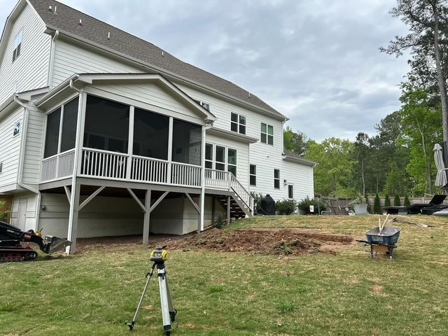 Backyard with raised screened porch on a white house, lawn, tripod, and wheelbarrow under cloudy sky