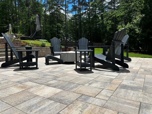 Outdoor stone patio with Adirondack chairs around a fire pit, surrounded by trees.