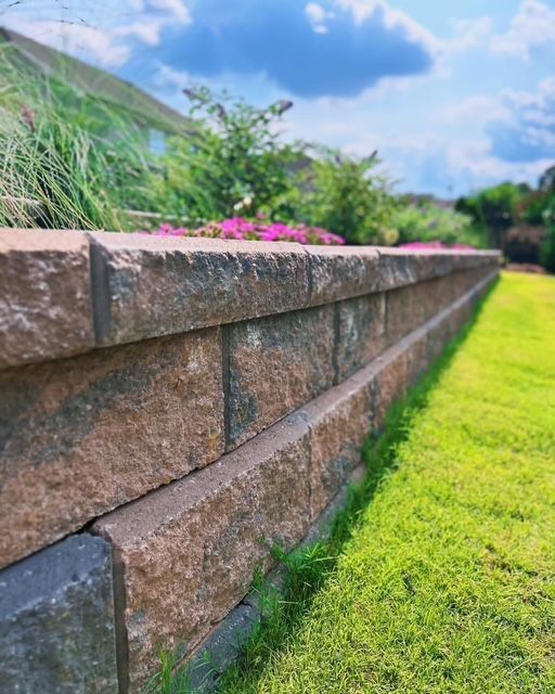 Sunlit stone retaining wall beside a green lawn with pink flowers and blue sky