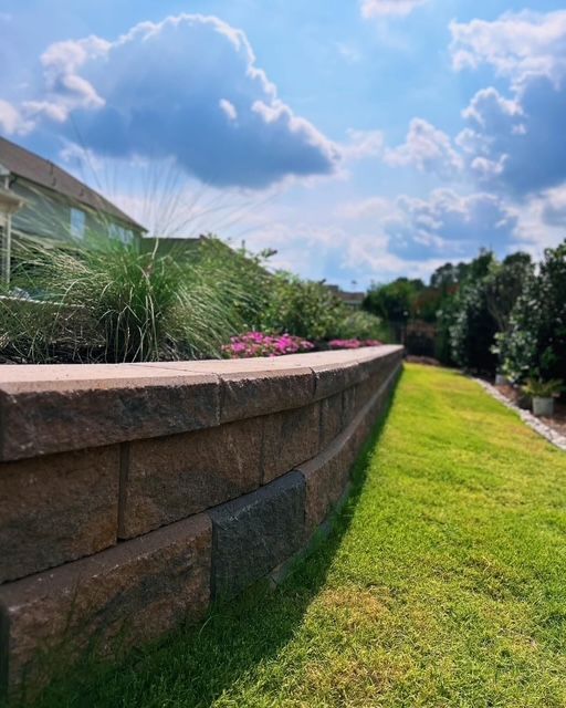 Curved stone retaining wall beside a green lawn and tall grasses under a bright blue sky with clouds