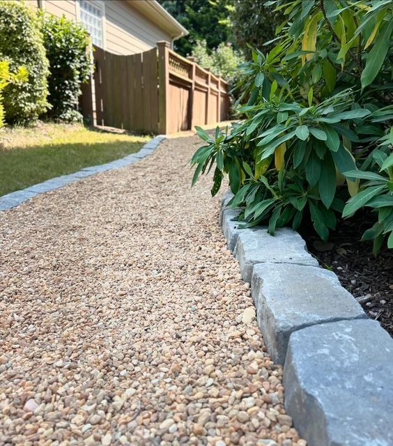 Gravel pathway beside a stone border and lush green shrubs along a house and fence