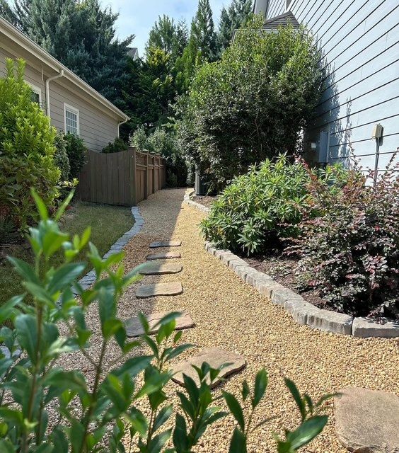 Narrow gravel side yard with stepping stones, shrubs, and tall trees between two houses