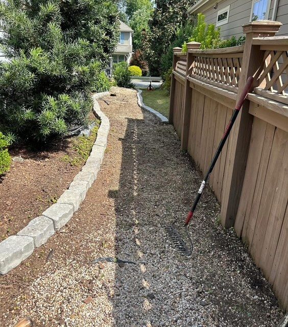 Gravel path beside a wooden fence and stone border in a landscaped yard, with a red-handled tool leaning on the fence