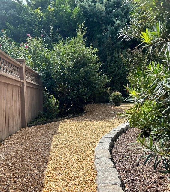 Curved gravel garden path beside a wooden fence and stone edging, surrounded by dense greenery