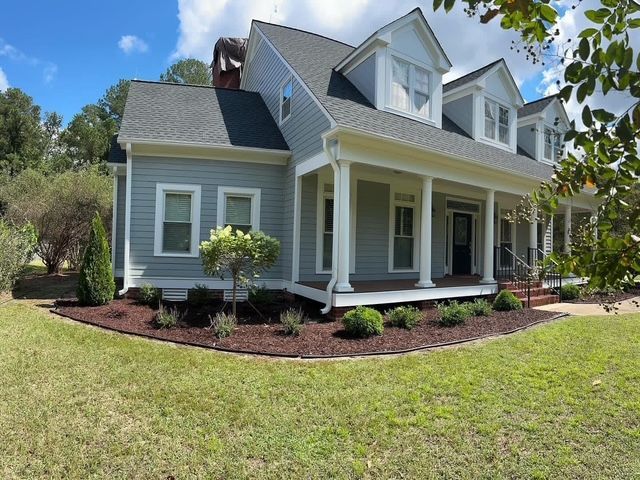 Gray house with white trim and front porch, surrounded by a green lawn and landscaped beds under a blue sky