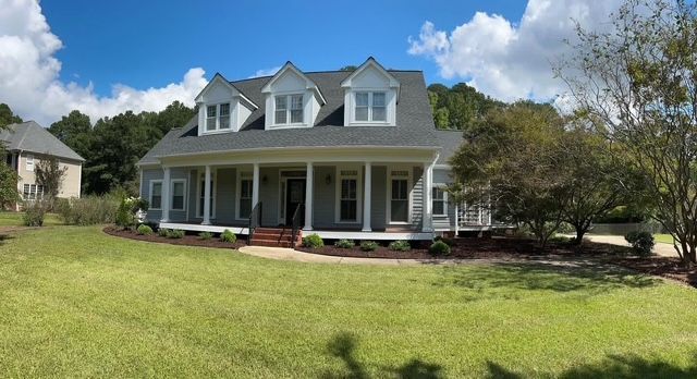 Large white farmhouse with dormer windows and a wraparound porch on a grassy lawn under a blue sky.