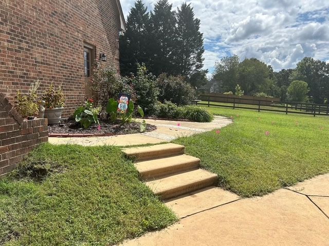 Brick house side yard with steps to a patio, landscaped garden, and green lawn under a partly cloudy sky
