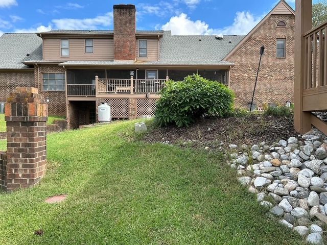 Backyard view of a brick house with a deck, chimney, green lawn, and stone-covered slope